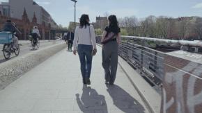 Das Szenenbild zeigt zwei Frauen von hinten, die nebeneinander über eine Brücke gehen.